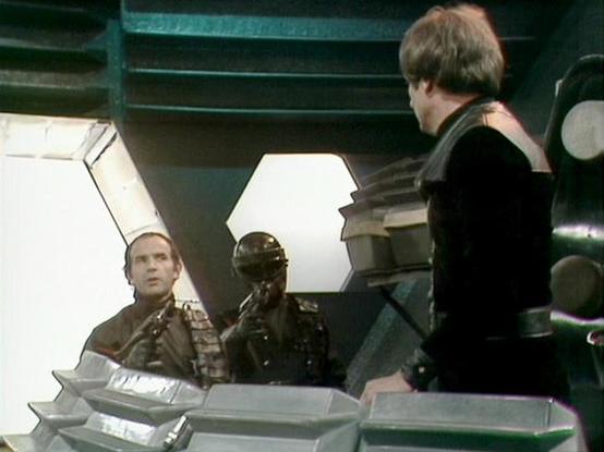 Claude 3.7 describes the image as: "This image appears to be from a science fiction television series, showing a scene set inside what looks like a spacecraft or futuristic control room. The image has the distinct visual style of older British sci-fi productions, with a somewhat minimalist set design featuring hexagonal lighting panels and metallic elements.

In the scene, several people are visible in what appears to be a tense moment. There's someone in dark clothing on the right side, while …