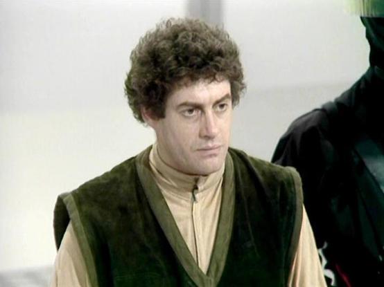 Claude Sonnet 4.0 describes the image as: "I can see this is a scene from a science fiction television series, showing a person with dark curly hair wearing what appears to be a green/olive colored vest or tunic over lighter colored clothing. The costume design has a futuristic or space-opera aesthetic typical of British sci-fi productions from the late 1970s or early 1980s. The setting appears to be indoors, possibly on a spaceship or futuristic facility set, with neutral colored walls or surf…