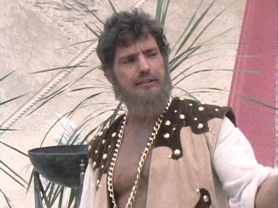Claude 3.7 describes the image as: "The image shows a person with curly hair and a beard wearing an ornate costume with a brown vest or shoulder piece decorated with white dots and gold trim over a white shirt. The setting appears to be outdoors with some palm fronds or similar vegetation visible in the background, suggesting a desert or tropical environment. The scene has a distinctive retro television production quality typical of British sci-fi shows from the late 1970s/early 1980s. The cost…