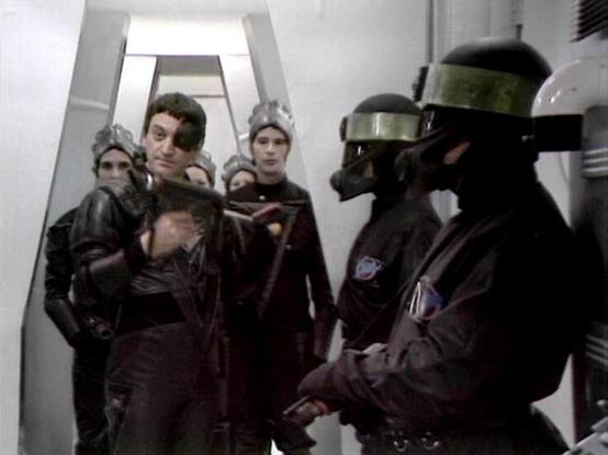 Claude 3.7 describes the image as: "This image appears to be from a science fiction television series, showing a scene in a stark, white corridor. Several people are visible wearing dark uniforms, with some wearing helmets or visors that obscure parts of their faces. The image has the distinctive low-budget, vintage sci-fi aesthetic typical of British television productions from the late 1970s/early 1980s.

The scene appears to depict some kind of confrontation in what looks like a spacecraft o…