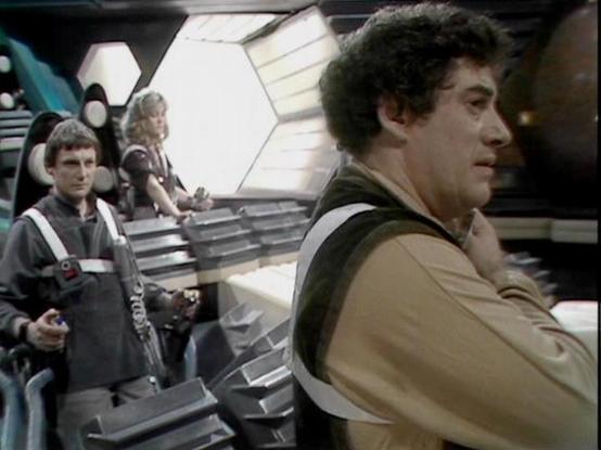 Claude 3.7 describes the image as: "This appears to be a still from a classic science fiction television series from the late 1970s/early 1980s. The image shows what appears to be the interior of a spacecraft or control room with a futuristic hexagonal design aesthetic typical of British sci-fi productions of that era.

The scene captures several performers in costume on what looks like a spaceship bridge or command center. The person on the right is wearing a light-colored outfit with dark tri…