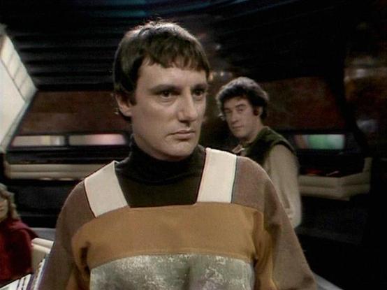 Claude Sonnet 4.0 describes the image as: "I can see this is a scene set aboard what appears to be a spacecraft, with the characteristic futuristic interior design typical of science fiction television from this era. The image shows two figures - one prominently in the foreground wearing a distinctive brown and beige uniform with geometric patterns, and another figure visible in the background. The setting appears to be the main area or control room of the ship, with the typical technological b…