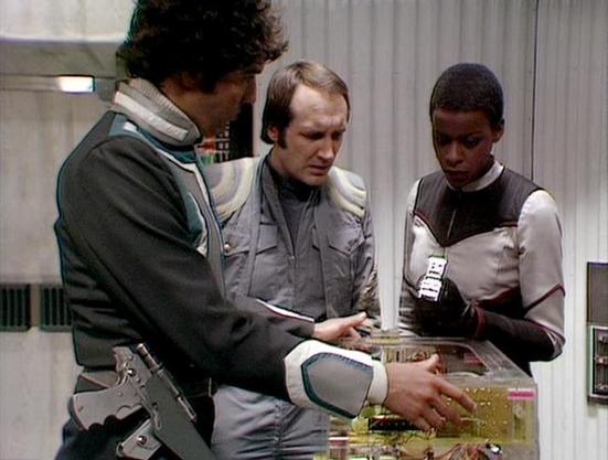 Claude 3.7 describes the image as: "The image shows three people in a sci-fi setting, looking at what appears to be some kind of control panel or electronic device. They're wearing distinctive futuristic costumes typical of classic British science fiction television from the late 1970s/early 1980s. 

The setting appears to be the interior of a spacecraft or space station, with utilitarian design elements visible in the background. The three individuals are examining something on a transparent o…