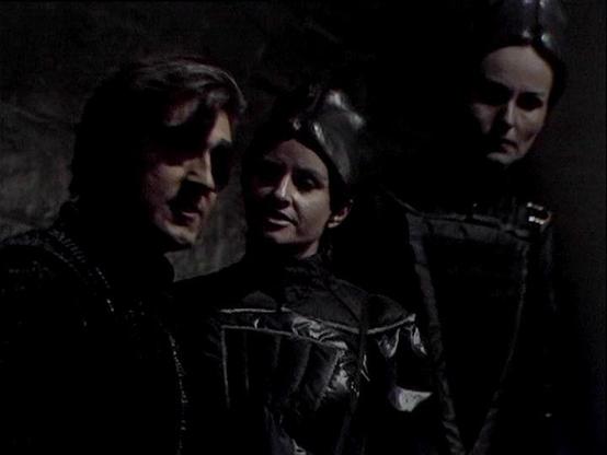 Claude 3.7 describes the image as: "This is a dark, atmospheric scene that appears to be from a science fiction television production from the late 1970s or early 1980s. The image shows three figures in what seems to be a dimly lit corridor or cave-like setting. 

The individuals are wearing distinctive costume pieces that include leather-like materials with geometric designs and structured elements typical of dystopian or space-oriented dramas of that era. The dramatic lighting creates strong …