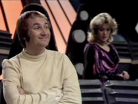 Claude 3.7 describes the image as: "The image shows a scene from a science fiction television production, featuring a person in a cream-colored turtleneck with arms crossed and a slight smile, appearing to be in some kind of futuristic set with geometric design elements. In the background, another person with blonde hair is wearing a purple/violet draped top. The setting appears to be a spacecraft or control room with distinctive lighting and angular structural elements typical of late 1970s/ea…