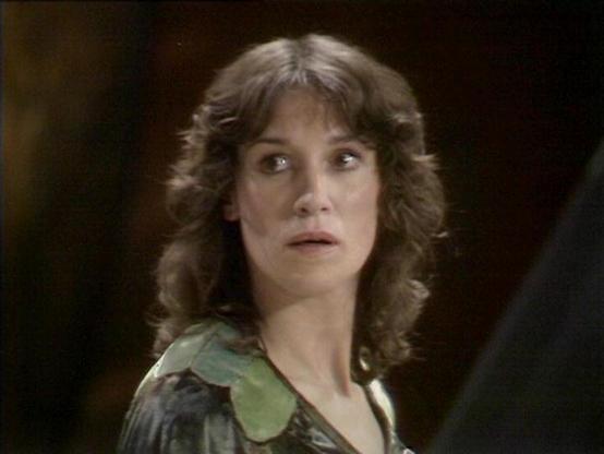 Claude Sonnet 4.0 describes the image as: "This image shows a character with shoulder-length wavy brown hair wearing what appears to be a green or olive-colored garment with distinctive collar details. The lighting creates a dramatic effect with a dark background, giving the scene a serious, tense atmosphere typical of science fiction television productions from this era. The character's expression appears concerned or alert, suggesting this may be from a moment of danger or uncertainty in the …