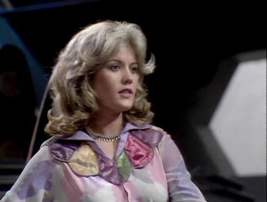 Claude Sonnet 4.0 describes the image as: "I can see this appears to be from a science fiction television production, likely from the late 1970s based on the styling and image quality. The scene shows someone with blonde, feathered hair wearing a colorful floral-patterned shirt or blouse in pink and purple tones, along with what appears to be a pearl necklace. The setting looks like it could be aboard a spacecraft or futuristic facility, with dark backgrounds and what might be technical equipme…