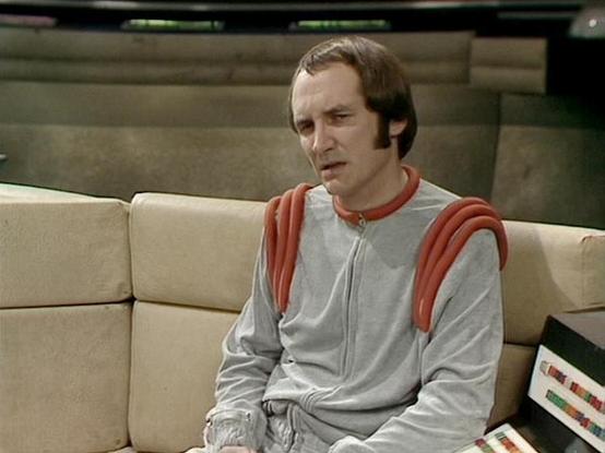 Claude 3.7 describes the image as: "The image shows a scene from a science fiction television series from the late 1970s/early 1980s. It features a person sitting on a light-colored couch or seating area that appears to be part of a spacecraft or futuristic setting. 

They're wearing a distinctive costume consisting of a light gray/silver jumpsuit with unusual red shoulder details. The costume has a minimalist, utilitarian design typical of sci-fi productions from that era.

The setting has the…