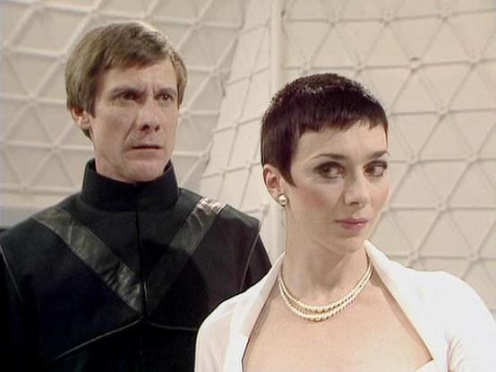 Claude Sonnet 4.0 describes the image as: "I can see two people in what appears to be a futuristic setting with white geometric paneled walls typical of science fiction productions. One person wears a dark military-style uniform with distinctive shoulder detailing, while the other is dressed in light-colored clothing with a pearl necklace and has very short dark hair. The staging and costumes suggest this is from a dramatic scene, likely taking place in some kind of command center or official f…