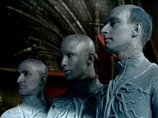 Claude Sonnet 4.0 describes the image as: "This image shows three humanoid androids or artificial beings with pale, bluish-gray skin and bald heads. They appear to be mechanical or cybernetic in nature, with visible seam lines or markings on their faces and necks that suggest artificial construction. The figures are positioned in profile, creating a striking visual composition. The setting appears to be some kind of industrial or spacecraft interior, with metallic structural elements visible in…