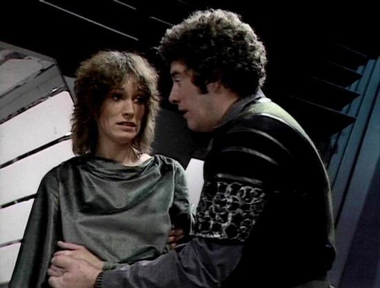 Claude Sonnet 4.0 describes the image as: "This appears to be a scene from a science fiction television series, showing two characters in what looks like a spacecraft or futuristic setting. The scene has a dramatic, intimate quality with the two figures positioned close together. One character is wearing what appears to be a green or grey garment, while the other is dressed in darker clothing. The lighting and set design are characteristic of British science fiction productions from the late 19…