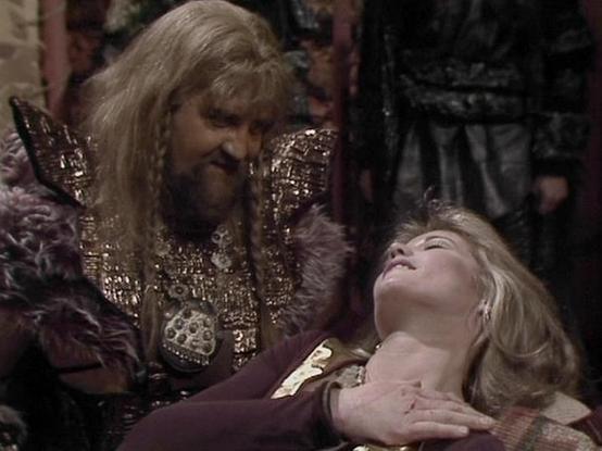 Claude Sonnet 4.0 describes the image as: "I can see this appears to be a dramatic scene with two characters in what looks like an ornate, medieval or fantasy-style setting. One figure is wearing elaborate, richly decorated clothing with metallic embellishments and appears to have long hair and facial hair. They are positioned over another character who appears to be lying down or reclining, with blonde hair and wearing what seems to be period costume. The scene has a theatrical, dramatic quali…