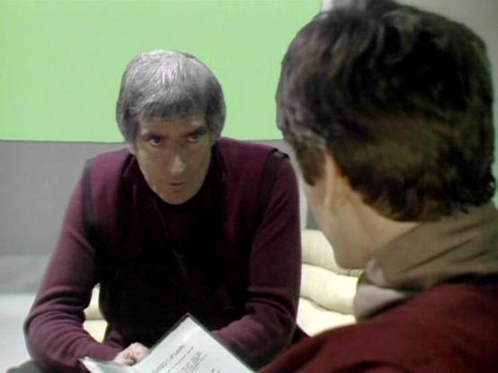 Claude 3.7 describes the image as: "This image shows a tense scene in what appears to be a minimalist, institutional setting with a green wall in the background. Two people are engaged in what looks like a serious conversation or interrogation. One person wearing a burgundy sweater with gray hair is leaning forward intently across what appears to be a table, while facing another person partially visible in the foreground. There appears to be some kind of document or paper between them that's be…