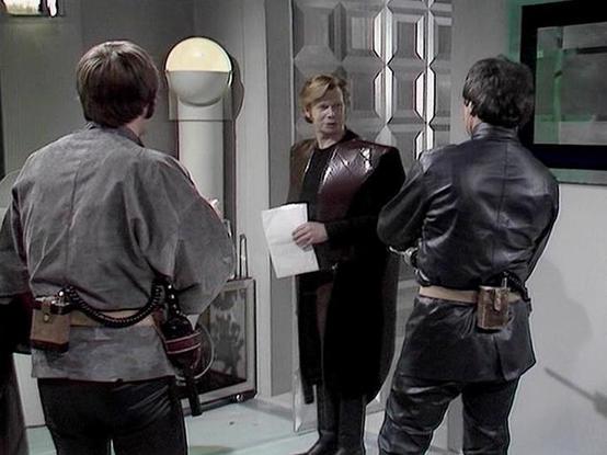 Claude 3.7 describes the image as: "This image shows a scene in a futuristic, sterile-looking spacecraft or space station interior with distinctive geometric wall panels. Three people are visible in what appears to be a tense confrontation. In the center, a person in a brown leather outfit holds what looks like a document or paper. They are facing two others - one wearing a grayish uniform with utility belts and equipment on the left, and another in dark clothing on the right. The setting has t…