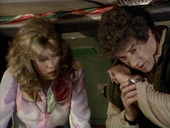 Claude 3.7 describes the image as: "The image shows a scene from a science fiction television series from the late 1970s or early 1980s. It appears to be set inside a spacecraft or control room, with some holiday decorations (candy canes) visible at the top of the frame.

Two people are shown in what seems to be a tense moment. On the left is a person with blonde, wavy hair wearing a pink jacket over a light-colored top. On the right is someone with curly dark hair wearing a beige sweater, spea…