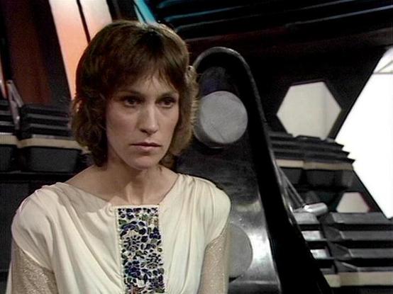 Claude 3.7 describes the image as: "The image shows a person with shoulder-length brown hair wearing a cream-colored outfit with a decorative floral pattern panel on the front. They appear to be in what looks like a futuristic or science fiction set, with geometric shapes and technological elements visible in the background. The setting has dark panels with some metallic components and distinctive lighting that creates a stark, dramatic atmosphere typical of classic science fiction television p…