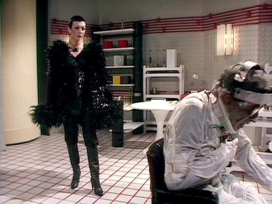 Claude Sonnet 4.0 describes the image as: "I can see this appears to be from a science fiction television series, showing a scene in what looks like a futuristic kitchen or laboratory setting with white tiled floors and modern equipment. There's a person in an elaborate black outfit with feathered or textured details standing prominently in the scene, while another figure in light-colored clothing appears to be crouched or kneeling near some equipment. The setting has a sterile, institutional f…