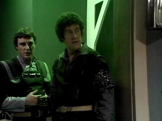 Claude Sonnet 4.0 describes the image as: "This appears to be a scene set in what looks like a spacecraft or futuristic facility interior, characterized by the distinctive green-tinted lighting and sleek metallic walls typical of science fiction productions from this era. Two men are visible in the frame - one wearing what appears to be a dark leather or fabric outfit with decorative elements, while the other is dressed in lighter colored clothing. The setting has the sterile, technological aes…