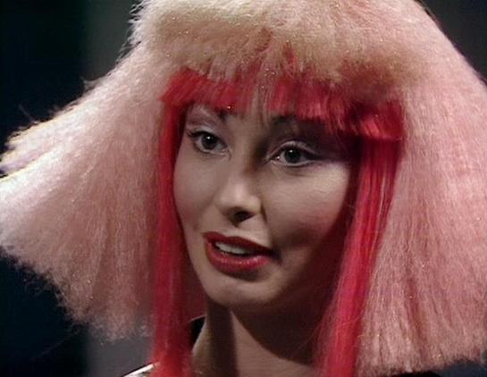 Claude 3.7 describes the image as: "The image shows a person with distinctive, dramatic hair styling featuring pink and red colors. The hairstyle is quite striking, with vibrant red bangs in the front and fluffy, voluminous light pink sections on the sides. They are wearing red lipstick and appear to be in a scene with dramatic lighting against a dark background.

This appears to be a costume design typical of science fiction productions from the late 1970s or early 1980s, with the bold, theatr…