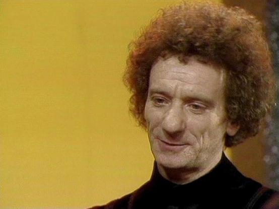 Claude Sonnet 4.0 describes the image as: "I can see this appears to be from a science fiction television production, likely from the late 1970s or early 1980s based on the visual style and production quality. The image shows someone with distinctive curly hair wearing what appears to be a dark colored garment, photographed against a yellowish background. The lighting and cinematography are characteristic of British television drama from that era. However, I'm not able to identify the specific …