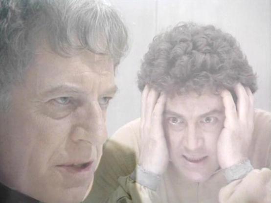 Claude 3.7 describes the image as: "The image shows a scene with a dramatic mood, featuring two people in what appears to be a tense or distressed situation. On the left is an older individual with a serious expression, while on the right is a younger person with curly hair who has their hands to their head in what seems to be a gesture of distress or confusion. 

The lighting has a muted, hazy quality that creates a dreamlike or psychological atmosphere, possibly suggesting this is from a scie…