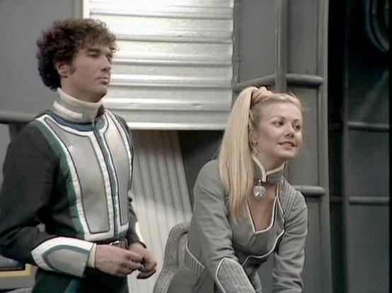 Claude 3.7 describes the image as: "This image shows a scene with two people in what appears to be a futuristic spaceship or station interior. They're wearing distinctive sci-fi costumes typical of late 1970s/early 1980s British television production. 

The person on the left wears a stylized outfit with green and white vertical stripes and a turtleneck, complemented by curly hair. The person on the right has blonde hair in a ponytail and wears a gray uniform with similar design elements and a …