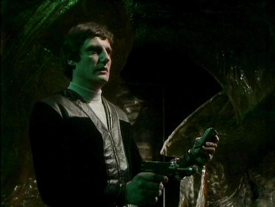Claude Sonnet 4.0 describes the image as: "This image shows a dramatic scene set in what appears to be a dark, underground or cave-like environment with rocky walls and atmospheric green lighting. The setting has a mysterious, possibly alien or futuristic quality typical of science fiction productions from this era. A character is positioned in the foreground, wearing dark clothing with what looks like a leather or similar material vest or jacket. The lighting creates strong shadows and gives t…