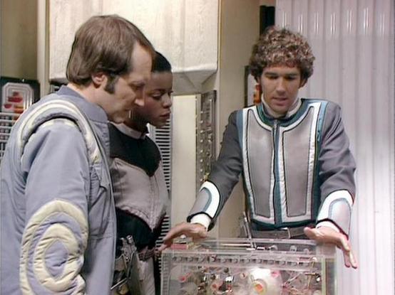 Claude Sonnet 4.0 describes the image as: "This scene appears to be set in a futuristic spacecraft or space station, with three characters gathered around what looks like a control panel or technical equipment. The setting has the characteristic sterile, technological aesthetic typical of science fiction productions from this era, with metallic surfaces and computer-like interfaces visible.

The characters are dressed in futuristic costumes - one wearing what appears to be a light-colored tunic…
