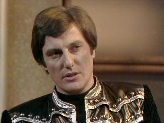 Claude 3.7 describes the image as: "The image shows a person in an elaborate costume that appears to be from a science fiction television production from the late 1970s or early 1980s. They are wearing a distinctive black outfit with ornate metallic silver and gold embellishments around the collar and shoulders, giving it a futuristic or ceremonial appearance.

The setting seems to be an interior scene with muted background colors, typical of studio television production of that era. The costum…