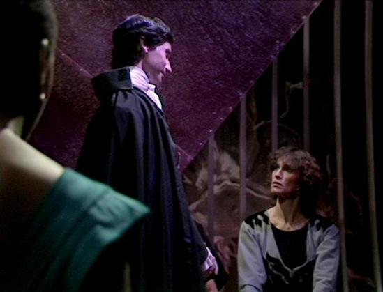 Claude Sonnet 4.0 describes the image as: "I can see this appears to be from a science fiction television series, showing what looks like a dramatic scene in a dungeon or prison-like setting with stone walls and metal bars. There are several people in the scene - one person wearing dark robes or a cloak is standing, while another person with curly hair appears to be seated or positioned lower. The lighting creates a moody, atmospheric effect typical of dramatic television productions from this …