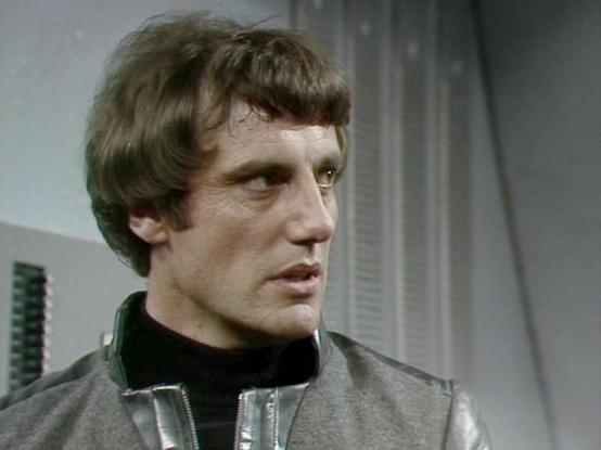 Claude 3.7 describes the image as: "This image appears to be from a science fiction television series from the late 1970s or early 1980s, based on the production quality and styling. The person in the image is wearing a distinctive gray leather or vinyl jacket with a black turtleneck underneath. They have dark, slightly tousled hair and are shown in what looks like a spacecraft or space station interior with metallic gray walls and control panels visible in the background. The lighting and set …