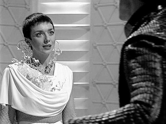 Claude 3.7 describes the image as: "This black and white image appears to be from a science fiction television production, featuring a futuristic setting with geometric wall patterns in the background. The scene shows a person in an elegant white draped garment with an ornate crystalline or transparent collar element. They are speaking to another figure partially visible on the right side of the frame, who appears to be wearing darker, textured clothing.

The stark lighting and minimalist set d…