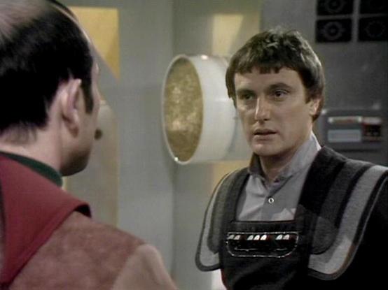 Claude Sonnet 4.0 describes the image as: "This scene shows two characters in conversation aboard a spacecraft. The setting appears to be inside a futuristic vessel with characteristic white and metallic interior design elements visible in the background, including circular porthole-like features. One character is wearing a dark leather outfit with decorative studs or metallic elements, while the other is dressed in what appears to be a reddish garment. The lighting and production design are ty…