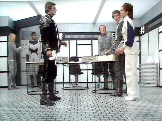 Claude 3.7 describes the image as: "This image shows a scene from a science fiction television series, set in what appears to be a futuristic control room or command center. Several people are gathered around a console with technical equipment. The setting features a clean, minimalist aesthetic typical of sci-fi productions from this era, with white walls, tiled flooring, and computer panels.

The individuals are wearing distinctive costumes - one in a black outfit with decorative elements stan…