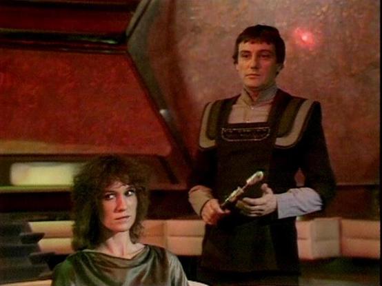 Claude 3.7 describes the image as: "This image appears to be a scene from a science fiction television series from the late 1970s or early 1980s. The setting features distinctive reddish-orange walls and futuristic design elements typical of sci-fi productions from that era.

The image shows two people in a spacecraft or space station interior. One person is standing, wearing a dark uniform with shoulder padding and holding what appears to be a futuristic device or weapon. The other person is s…
