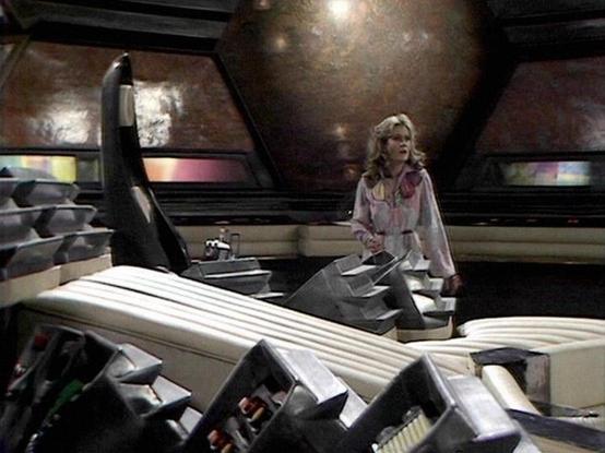 Claude Sonnet 4.0 describes the image as: "I can see this is a science fiction setting with futuristic interior design featuring curved walls, metallic surfaces, and what appears to be a spacecraft or space station environment. There's sophisticated control equipment and seating visible in the foreground. The set design has the distinctive aesthetic of 1970s British science fiction television, with smooth, angular surfaces and muted lighting that creates an atmospheric, somewhat sterile spacesh…
