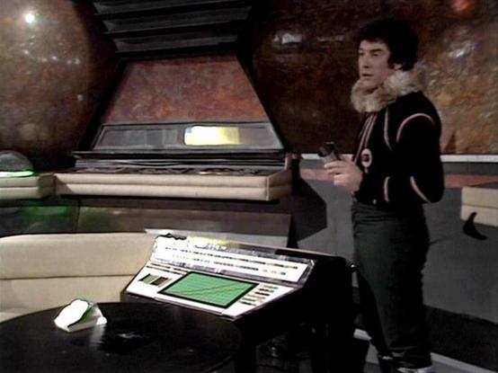 Claude 3.7 describes the image as: "This image shows a scene from what appears to be a science fiction television series from the late 1970s or early 1980s, based on the production style and set design. The scene takes place in what looks like a spacecraft or control room with distinctive textured copper/brown walls and futuristic control panels.

In the foreground is a console with a green display screen and various controls. A person is standing in the set wearing a dark outfit with a light-c…