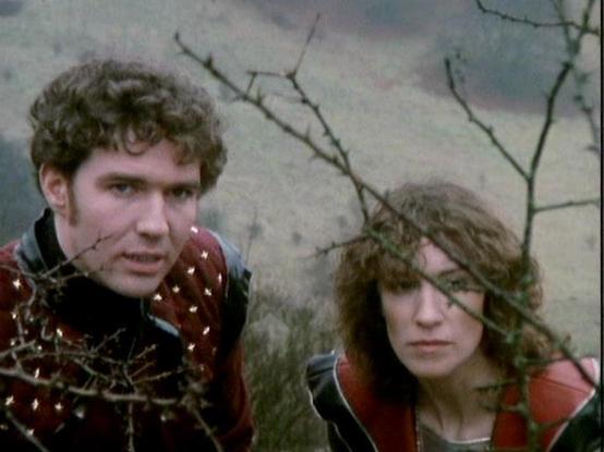 Claude Sonnet 4.0 describes the image as: "I can see two people in what appears to be a science fiction setting, likely from a dramatic scene. They're positioned against what looks like a rocky or cave-like background with bare branches visible, creating an outdoor or alien landscape atmosphere. Both individuals are wearing what appears to be futuristic or period costume clothing in earth tones. The lighting and film quality suggests this is from a television production of an earlier era. The s…