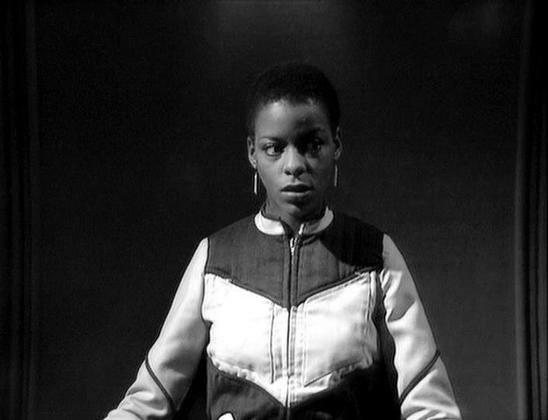 Claude 3.7 describes the image as: "This is a black and white image from a dramatic television series from the late 1970s/early 1980s British science fiction era. The frame shows a person wearing a distinctive two-tone outfit with a white and dark color-blocked design featuring a zippered front. The person is wearing small hoop earrings and has a serious, intense expression against a dark background, creating a stark contrast typical of the dramatic lighting used in this production.

The minima…