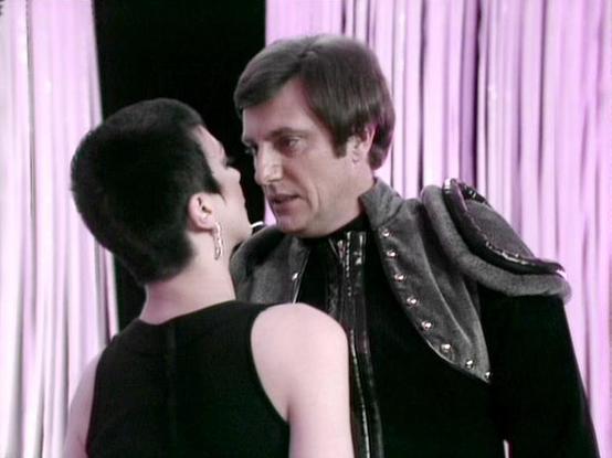 Claude Sonnet 4.0 describes the image as: "I can see this appears to be from a science fiction television series, showing two characters in what looks like an intimate or tense dramatic moment. One person is wearing a black outfit while the other is dressed in dark clothing with decorative metallic studing or armor-like details. The setting appears to be an interior space with curtains or draping visible in the background, lit with purple/pink lighting that creates a dramatic atmosphere. The sc…