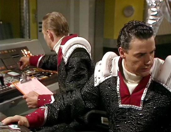 Claude 3.7 describes the image as: "The image shows two individuals in a science fiction setting, wearing distinctive costume uniforms with black sparkly material, white shoulder pads, and red accents. They appear to be operating some kind of control panel or spacecraft console with various buttons and instruments. The costume design has a retro futuristic aesthetic typical of classic British sci-fi productions from the late 1970s/early 1980s. 

The setting appears to be some sort of command ce…