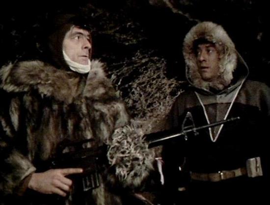 Claude 3.7 describes the image as: "The image shows two people in what appears to be a cold, wintery setting, possibly a cave or rocky environment. Both individuals are wearing heavy fur-lined winter clothing and parkas with hoods designed for extreme cold weather conditions. One person on the left appears to be wearing a bandage or wrapping around their neck/jaw area, while both are equipped with weapons. The dark, rocky background suggests they may be in a hostile environment, perhaps during …