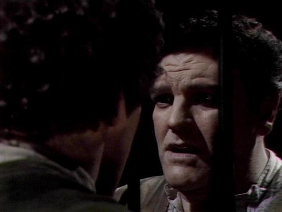 Claude 3.7 describes the image as: "The image shows a tense confrontation scene from a dark, atmospheric science fiction production. It appears to be from a dramatic moment where two people are having an intense exchange. The lighting is low-key and moody, with one person partially visible in the foreground from behind, while another person with a concerned or troubled expression is visible in the light. They're wearing what appears to be simple clothing typical of the show's aesthetic.

The se…