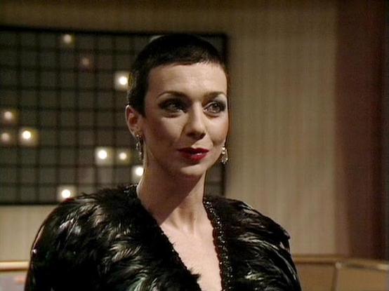 Claude Sonnet 4.0 describes the image as: "I can see this is from a science fiction television series, showing an elegantly dressed character in what appears to be a luxurious interior setting. The person is wearing a dramatic black outfit with feathered or textured shoulders, and has striking makeup with bold red lipstick. Their hair is styled in a sleek, short fashion typical of futuristic or avant-garde looks. The background features warm lighting and what appears to be decorative elements, …