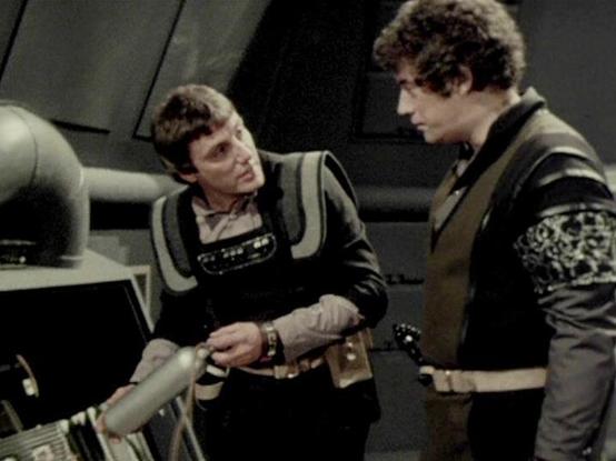 Claude Sonnet 4.0 describes the image as: "This appears to be a scene set aboard a spacecraft, likely the Liberator, showing two crew members in a technical or engineering area. The setting features the show's characteristic futuristic interior design with metallic surfaces and technological equipment. One character is leaning over what appears to be some kind of control panel or technical apparatus, while another stands nearby observing. Both are wearing the dark, military-style clothing typic…