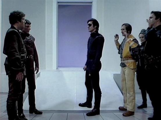 Claude Sonnet 4.0 describes the image as: "This appears to be a scene from a science fiction television series set in a sterile, white-walled futuristic environment. Several characters are gathered in what looks like a spaceship or space station corridor, with a doorway or window showing a purple/violet glow in the background. The characters are wearing various costumes typical of 1970s/80s sci-fi productions - including dark leather or fabric outfits and more casual clothing. The setting and c…