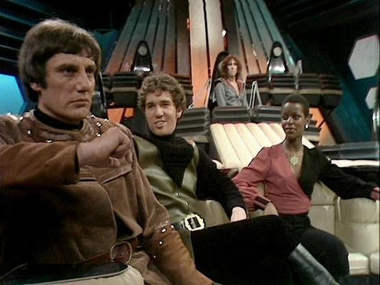 Claude 3.7 describes the image as: "This image shows a scene from a sci-fi television series set in what appears to be a spacecraft control room or command center. The futuristic set features distinctive ribbed wall panels and technical equipment typical of 1970s/80s British television science fiction production design.

Four performers are shown in the spacecraft interior, seated in what looks like a command area. One person wearing a brown leather-like outfit with buckles sits prominently in …