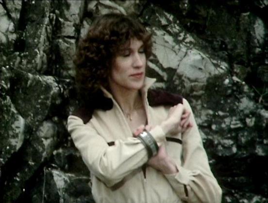 Claude 3.7 describes the image as: "The image shows a person with curly dark hair wearing a cream-colored outfit with dark accents against a rocky backdrop. The clothing style and the film quality suggest this is from a classic science fiction television production, likely from the late 1970s or early 1980s based on the aesthetic. 

The setting appears to be an outdoor location with stone or rock formations creating a somewhat stark, possibly alien landscape. The costume has a distinctive desig…