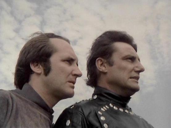 Claude 3.7 describes the image as: "The image shows two people in profile against a cloudy sky background. The composition has them positioned side by side, both looking toward the same direction. The person on the left wears what appears to be a gray or brown outfit, while the person on the right is dressed in a distinctive black leather costume with metallic studs or embellishments along the collar.

The styling and aesthetic suggests this is from a vintage science fiction production, likely …