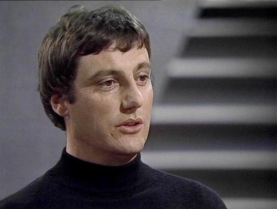 Claude 3.7 describes the image as: "The image shows a person in what appears to be a scene from a television production, likely from the late 1970s or early 1980s based on the visual style and quality. They are wearing a black turtleneck sweater and have short dark hair. The background is minimalist and gray, with what looks like stairs or stepped architecture visible behind them.

The lighting and cinematography have that distinctive quality common in British sci-fi television productions of t…
